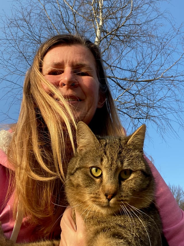 Maja Berden Zrimec outdoors in winter sunlight, holding her tabby cat, with a bare birch tree behind them.